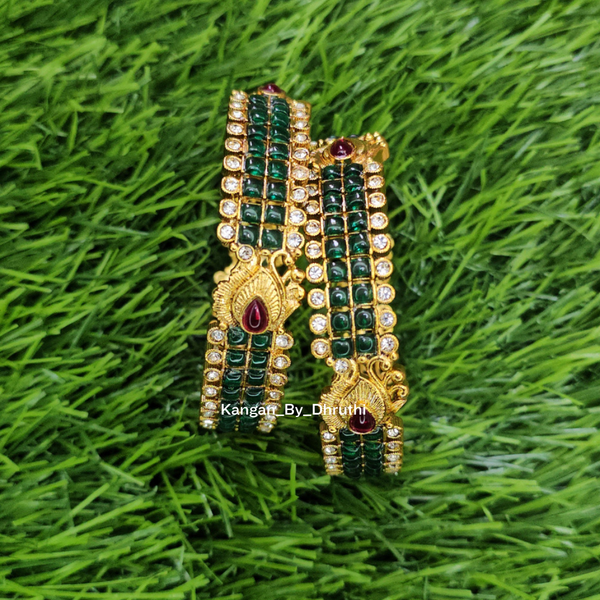 A pair of gold-plated traditional Indian bangles decorated with green stone stripes and red teardrop gemstones, displayed on a green grass background.
Green Kemp Micro-Gold Plated Peacock Bangles