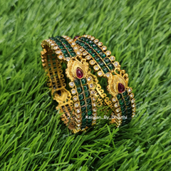 A set of gold-plated Indian-style bangles decorated with green stone-encrusted rows and red teardrop gemstones, displayed on a green grass background. Green Kemp Micro-Gold Plated Peacock Bangles