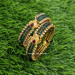 A set of traditional Indian bangles featuring gold-plated metal with green stone-encrusted rows and red oval gemstone accents, displayed on a green grass background. Green Kemp Micro-Gold Plated Peacock Bangles