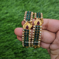 A pair of gold-plated bangles decorated with green and white stones, held in a hand against a green grass background. Green Kemp Micro-Gold Plated Peacock Bangles