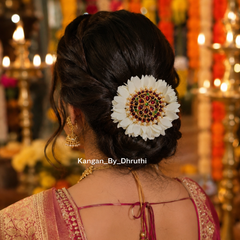 A woman shown from behind wearing a white artificial flower hair accessory with a jeweled center, styled in her hair inside a warmly lit ceremonial setting.
