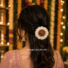 A woman shown from the back wearing a half-updo hairstyle with a large white artificial flower hair accessory featuring a brown patterned center, a pink embroidered blouse, and gold earrings, set against a festive background with hanging garlands and warm lights.