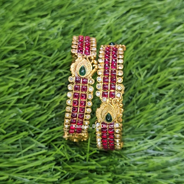 A white daisy-style bangle with a gold-toned center set with red, green, and white stones, placed on a green grass-like textured background. Ruby-Pink Kemp Micro-Gold Plated Peacock Temple Bangles