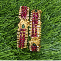 A pair of gold-plated traditional Indian Kundan bangles decorated with red stones, clear stones, and green teardrop accents, displayed on a green grass background. Ruby-Pink Kemp Micro-Gold Plated Peacock Temple Bangles