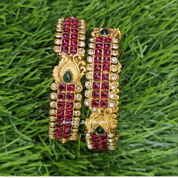 A pair of gold-plated traditional Indian Kundan bangles decorated with red stones, clear stones, and green teardrop accents, displayed on a green grass background. Ruby-Pink Kemp Micro-Gold Plated Peacock Temple Bangles
