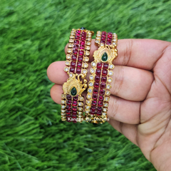 A pair of gold-plated multi-strand bangles decorated with deep red stones and green teardrop-shaped center stones, held in a person's hand against a green grass background. Ruby-Pink Kemp Micro-Gold Plated Peacock Temple Bangles