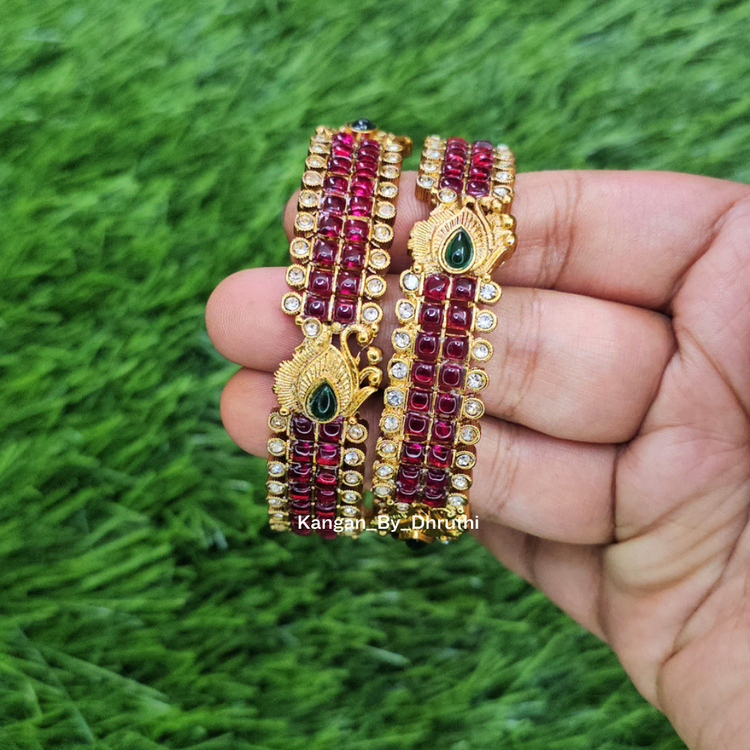 A pair of gold-plated multi-strand bangles decorated with deep red stones and green teardrop-shaped center stones, held in a person's hand against a green grass background. Ruby-Pink Kemp Micro-Gold Plated Peacock Temple Bangles