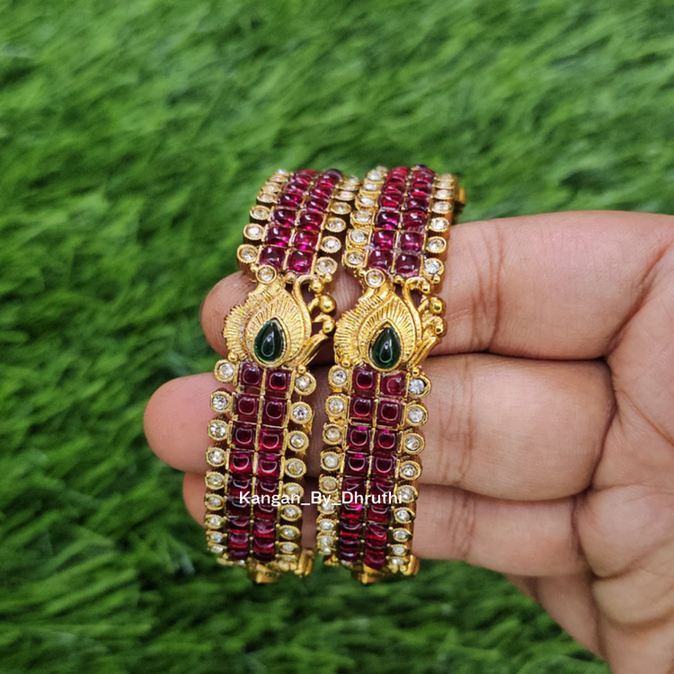 A hand holding a set of gold-plated bangles decorated with rows of deep red and clear rhinestones, featuring green stone accents at the center, set against a green grass background. Ruby-Pink Kemp Micro-Gold Plated Peacock Temple Bangles
