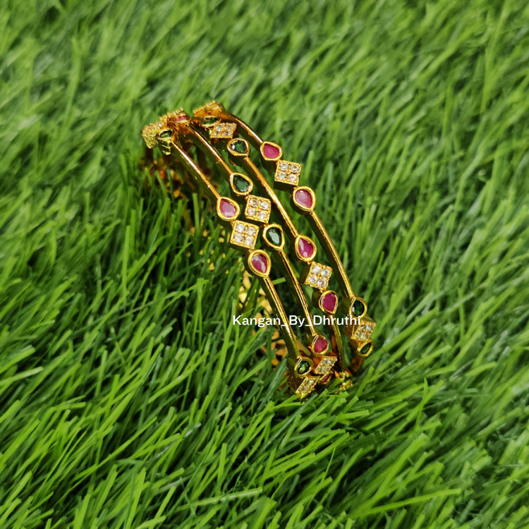 A gold-plated multi-strand bangle bracelet decorated with red, green, and clear stones, resting on a background of green grass.
Ruby Pink & Emerald Green Micro-Gold Plated Temple Bangles
