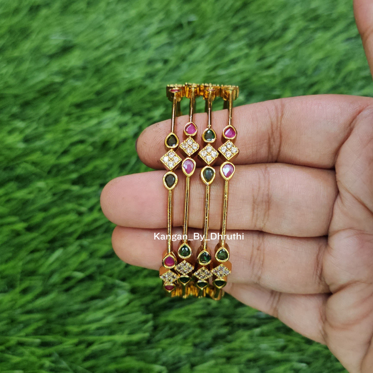 A hand holding four gold-plated bangles decorated with green, pink, and clear stones, set against a green grass background. 
Ruby Pink & Emerald Green Micro-Gold Plated Temple Bangles