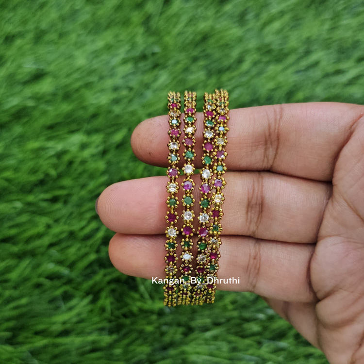 A hand holding three gold-plated bangles decorated with small pink, green, and white floral beads, set against a blurred green grass background. Ruby Pink & Green Antic Finish Bangles