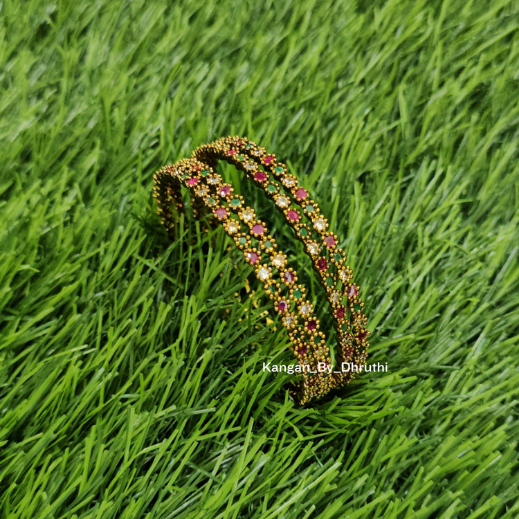 A gold-plated multi-strand bangle bracelet decorated with pink and green floral beadwork, resting on a green grass background. Ruby Pink & Green Antic Finish Bangles
