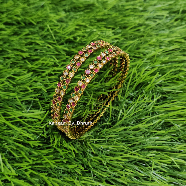 A gold-plated beaded double bangle bracelet decorated with pink and white stones, displayed on a green grass background. Ruby Pink & Green Antic Finish Bangles