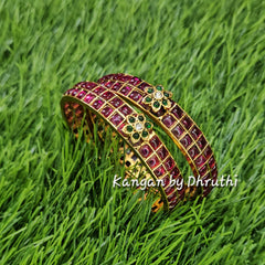 A gold-plated bangle bracelet with red and green stone inlay, resting on a green grass background. A watermark 'Kangan by Dhruchi' is visible on the right side.Ruby Pink & Green Floral Motif Gold Imitation Bangles