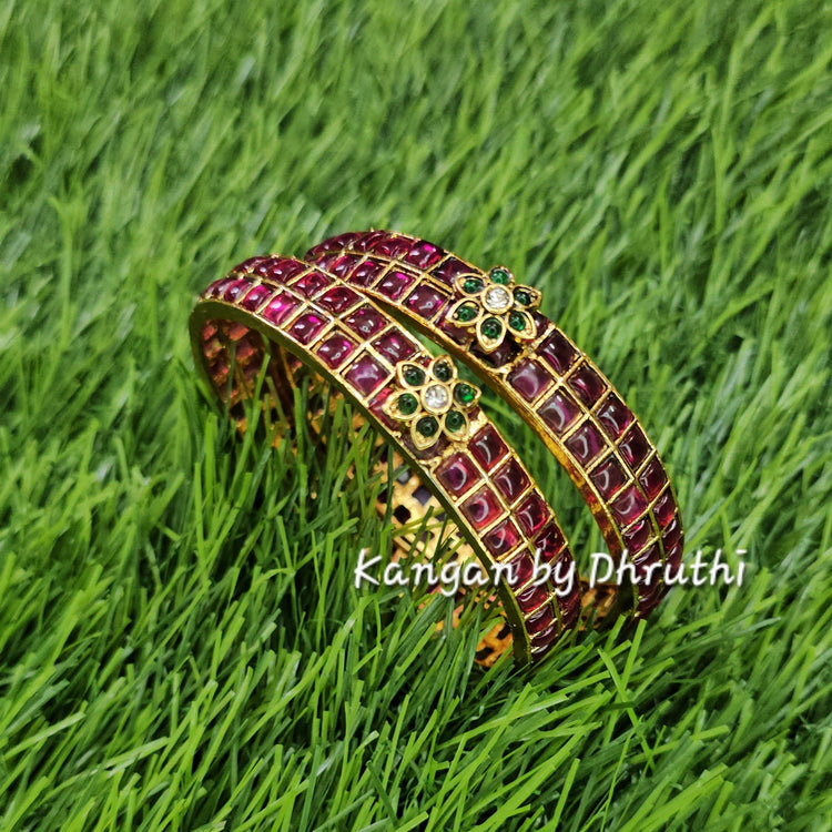 A gold-plated bangle bracelet with red and green stone inlay, resting on a green grass background. A watermark 'Kangan by Dhruchi' is visible on the right side.Ruby Pink & Green Floral Motif Gold Imitation Bangles