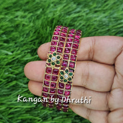 A hand holding a multi-strand bracelet with pinkish-red stones and a central green and gold floral motif, set against a green grass background.Ruby Pink & Green Floral Motif Gold Imitation Bangles