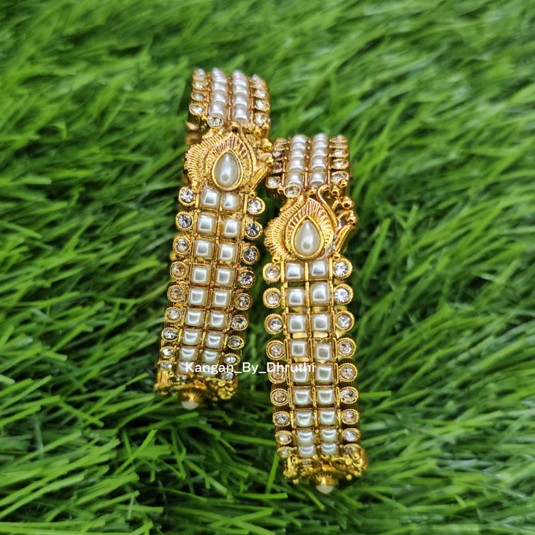 A pair of gold-plated traditional Indian bangles decorated with white faux pearls and small clear stones, displayed on a green grass-like textured background. White Pearl Micro-Gold Plated Peacock Bangles