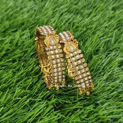 Two ornate gold-toned bangles decorated with white stones and central pearl-like accents, displayed on a green grass-like surface. White Pearl Micro-Gold Plated Peacock Bangles