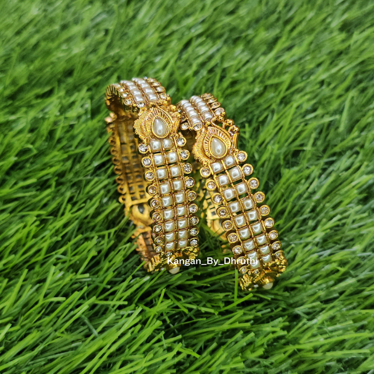 Two ornate gold-toned bangles decorated with white stones and central pearl-like accents, displayed on a green grass-like surface. White Pearl Micro-Gold Plated Peacock Bangles