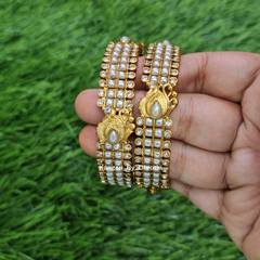 A hand holding two gold-plated multi-strand bangles decorated with white faux pearls and small clear stones, set against a green grass background. White Pearl Micro-Gold Plated Peacock Bangles