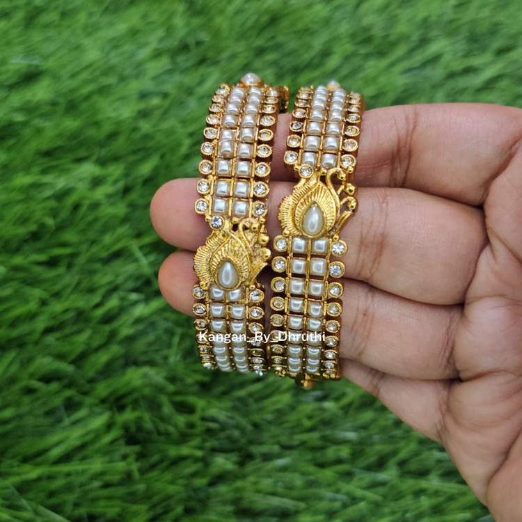 A hand holding two gold-plated multi-strand bangles decorated with white faux pearls and small clear stones, set against a green grass background. White Pearl Micro-Gold Plated Peacock Bangles